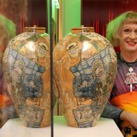 A woman standing between two mirrored ceramic urns in a red-curtained room, from 'Queer Urns and Quilts'