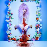 Surreal image of a figure hanging upside down inside a floral wreath frame, with red liquid dripping below