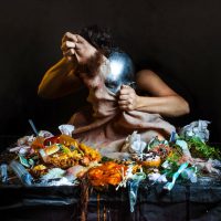 A dramatic photograph of a figure surrounded by scattered food and flowers, holding a glowing orb to their face, from 'The Horror of Sarah Sitkin'