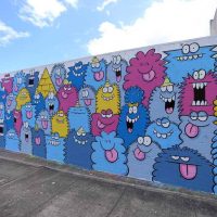 An outdoor mural covered in rows of colorful cartoon monster faces, titled 'Little Monsters at POW! WOW!'