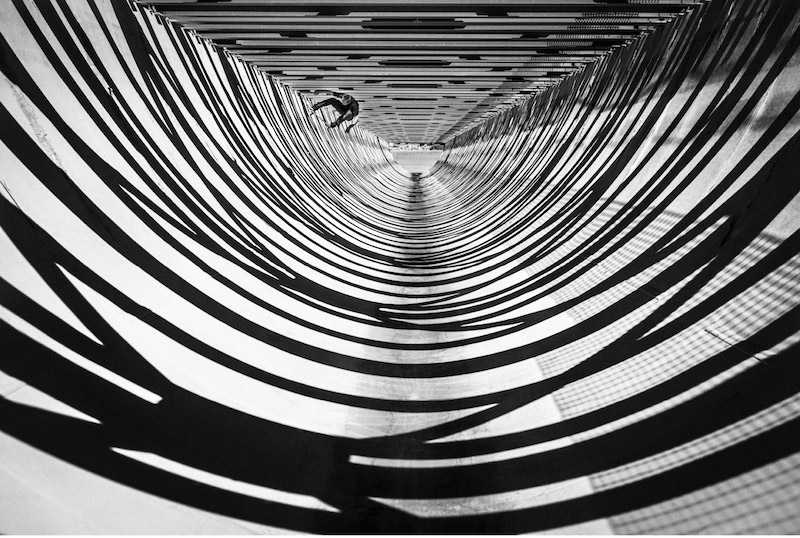 A black-and-white abstract image of swirling, concentric tunnel-like lines, titled 'Rolling Through the Shadows'
