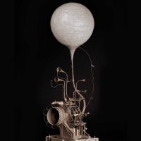 Photograph of a small mechanical sculpture with a white balloon against a black background
