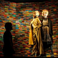 Two robed figures standing before a wall of colorful vertical stripes, with one figure in silhouette, by Nick Georgiou