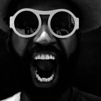 Black-and-white photograph of a person wearing round glasses and a hat, shouting
