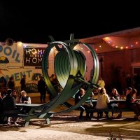 Nighttime view of a large twisted green ribbon sculpture by Michael Beitz with people gathered at picnic tables under string lights