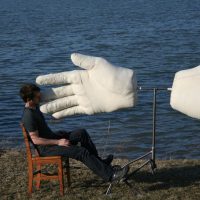Surreal photograph of oversized white glove sculptures reaching toward a man seated by a lake, by Michael Beitz