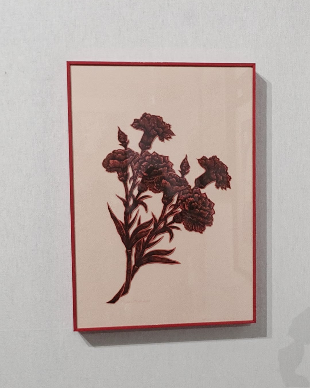 Framed work showing a bouquet of carnations in dark red on pale pink paper, in a red frame on a grey wall