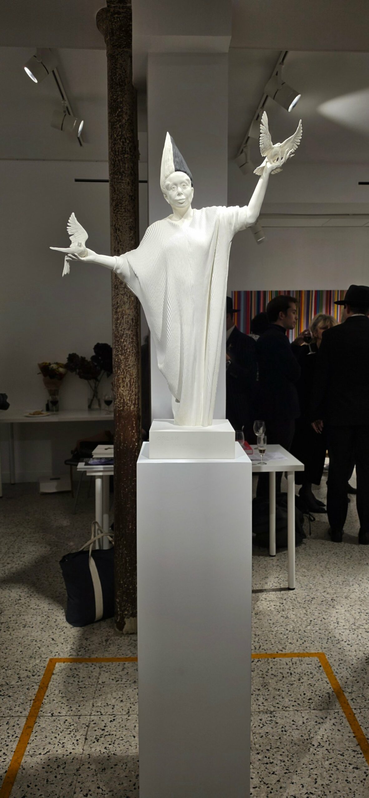 A white sculpture of a figure with a tall conical headpiece and a pleated robe, holding a dove on each outstretched hand, on a white plinth in the gallery.