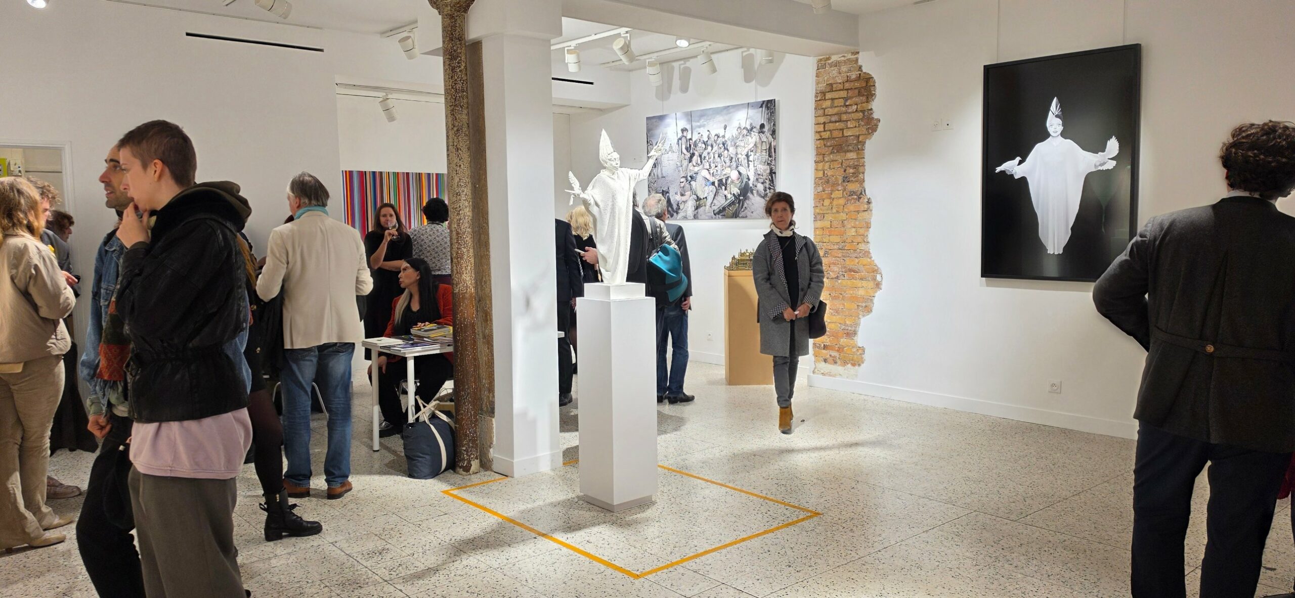 Wide view of the opening: the white dove-bearing sculpture on a plinth, a large photograph of soldiers around a table, and a framed black-and-white photograph of a figure with doves on the right wall, with visitors.