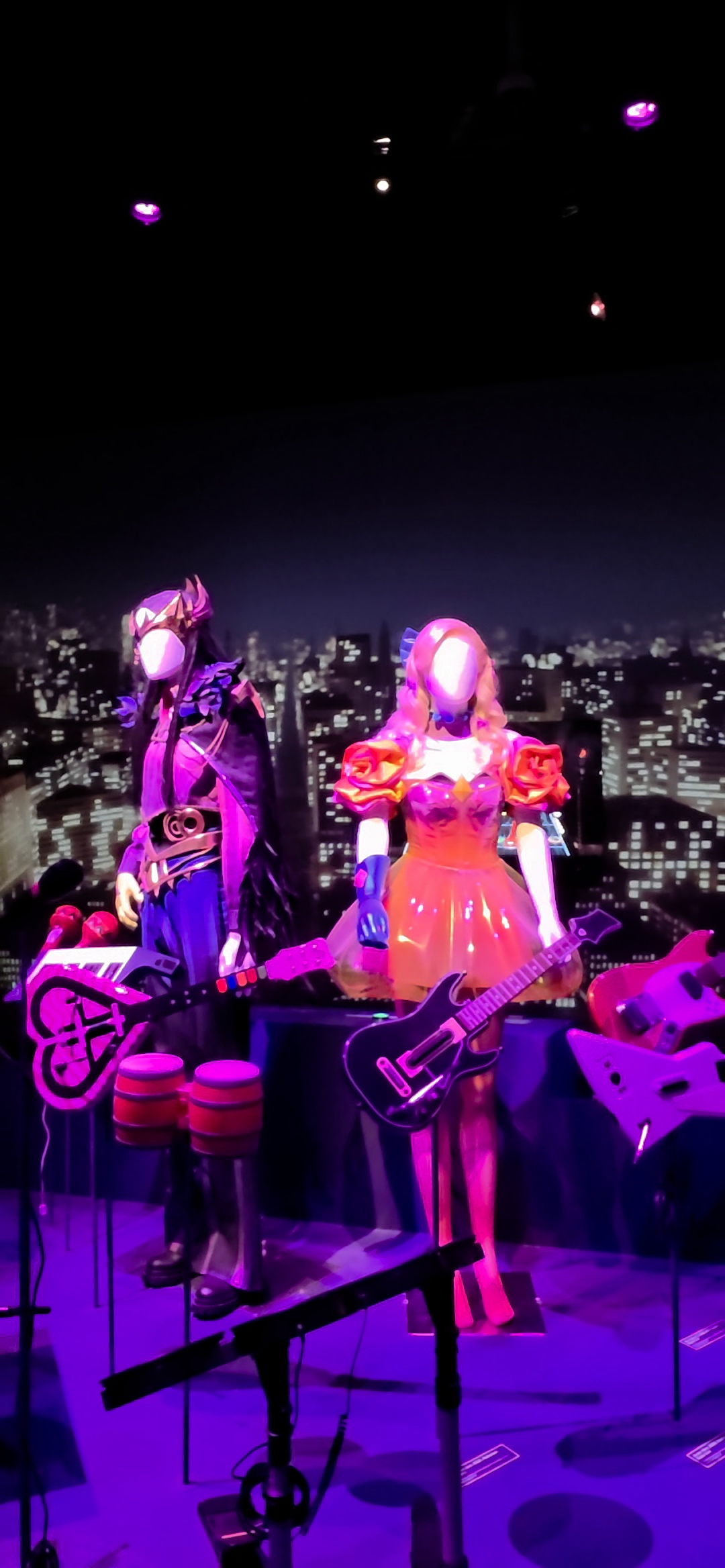 Deux mannequins en costumes de scène parmi des manettes en forme de guitare et des tambours, devant un horizon de ville nocturne