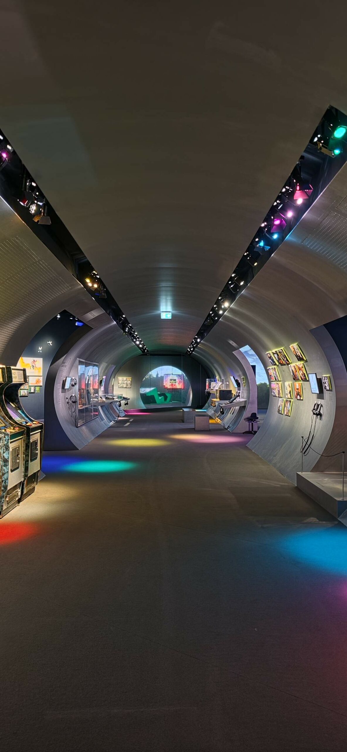 Un tunnel courbe en métal dans l’exposition, éclairé par des projecteurs colorés, avec des bornes d’arcade à gauche et des œuvres encadrées à droite