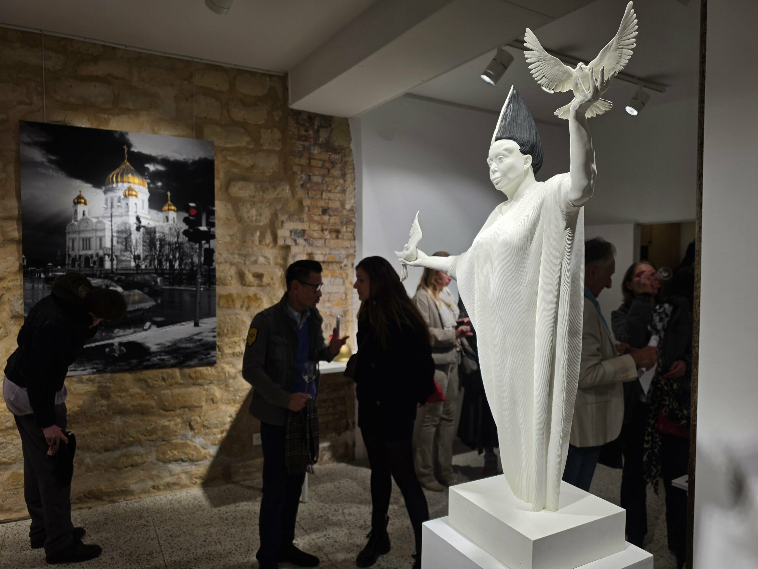 Sculpture blanche d’un personnage en robe plissée et haute coiffe conique, bras écartés, une colombe sur chaque main, à côté d’une photographie en noir et blanc d’une cathédrale aux dômes dorés ; des visiteurs à l’arrière-plan.